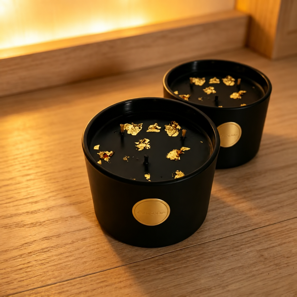 Two black and gold three-wick soy candles on a wooden shelf in warm light